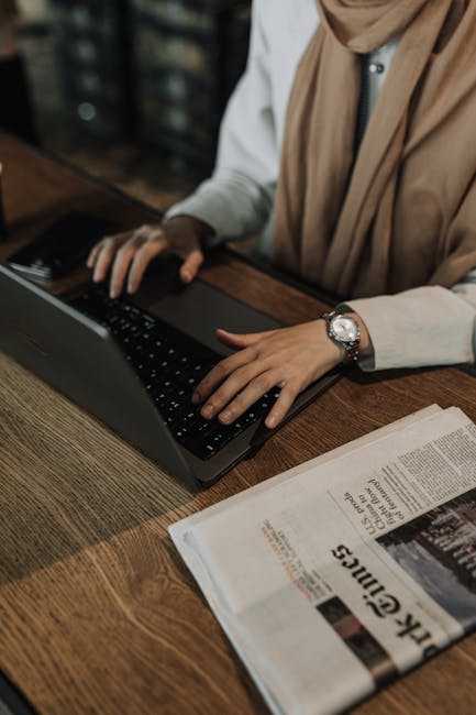 Woman working on a laptop at a wooden desk with a newspaper beside her.