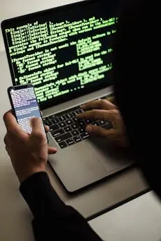 A hooded figure engaged in hacking using a laptop and smartphone in low light.