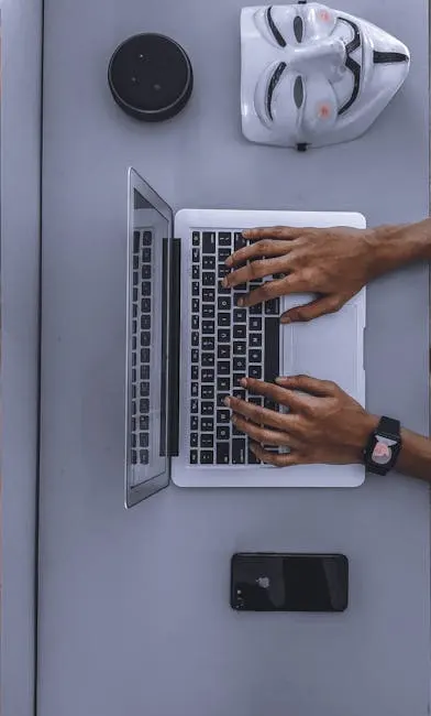 Top view of hands typing on a laptop beside a mask and smartphone.